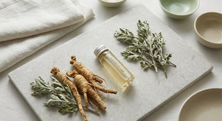 A close-up flat-lay of ginseng roots, dried mugwort sprigs, and a glass vessel of clear bamboo sap arranged on a stone surface in soft natural light.
