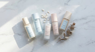 A frost-dusted marble flat lay of minimalist skincare bottles arranged in an arc with scattered white capsules and a dried botanical sprig, lit by cool diffused natural light in icy blue and creamy white tones.