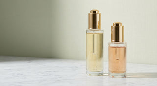 Two glass dropper bottles containing skincare serums in amber and gold tones resting on a marble surface in soft natural light.